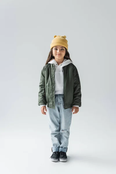 Full length of adorable girl in trendy autumnal outfit smiling and standing on grey - foto de stock
