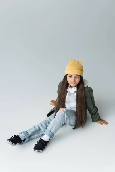 High angle view of adorable girl in trendy autumnal outfit and beanie hat sitting on grey - foto de stock