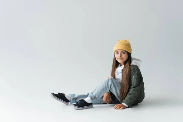 Full length of adorable girl in trendy autumnal outfit and beanie hat sitting on grey — Photo de stock