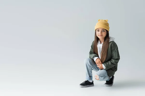 Повна довжина радісної і милої дівчини в модному автономному вбранні і капелюсі бані на сірому — стокове фото