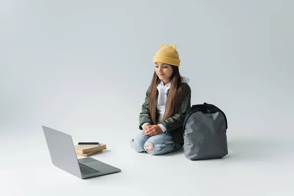 Full length of adorable girl in trendy autumnal outfit looking at laptop while sitting near smartphone on books and backpack on grey — Stock Photo