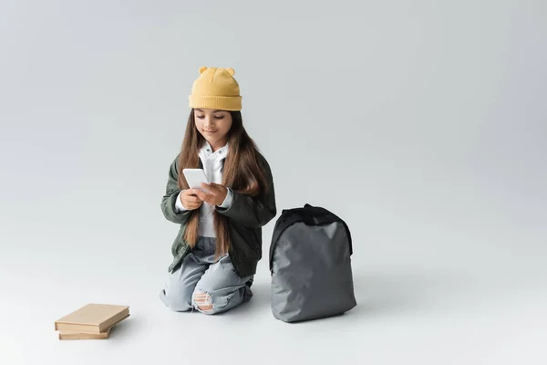Full length of adorable girl in trendy autumnal outfit using smartphone while sitting near books and backpack on grey — Photo de stock