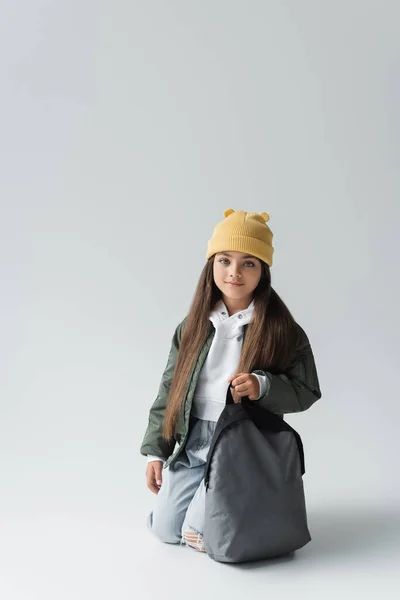 Full length of cheerful kid in trendy autumnal outfit and beanie hat holding backpack on grey - foto de stock