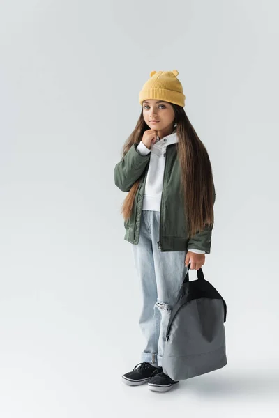 Full length of adorable girl in trendy autumnal outfit and beanie hat holding backpack on grey — Stock Photo