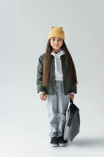 Full length of smiling and cute girl in trendy autumnal outfit holding backpack on grey — стоковое фото