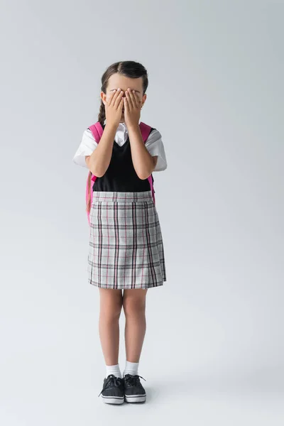 Повна довжина дівчини в шкільній формі покриває обличчя, стоячи на сірому — стокове фото