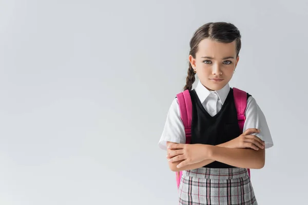 Sad schoolgirl with backpack standing with crossed arms isolated on grey — Stock Photo