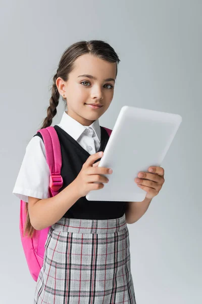 Щаслива і мила школярка в уніформі тримає цифровий планшет, посміхаючись ізольовано на сірому — стокове фото