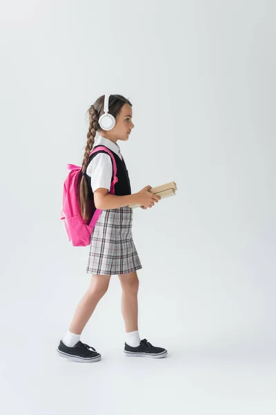 Side view of girl in school uniform and wireless headphones walking with books on grey — стоковое фото