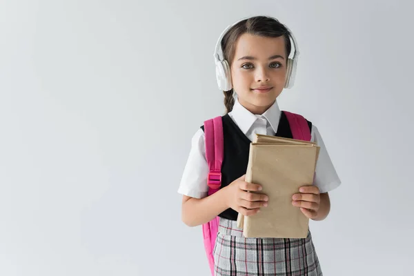 Дівчина в шкільній формі і бездротові навушники тримають книги ізольовані на сірому — стокове фото