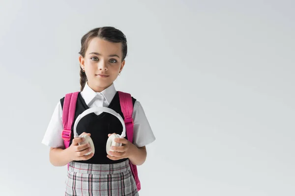 Schoolgirl in uniform standing with backpack and holding wireless headphones isolated on grey — Foto stock