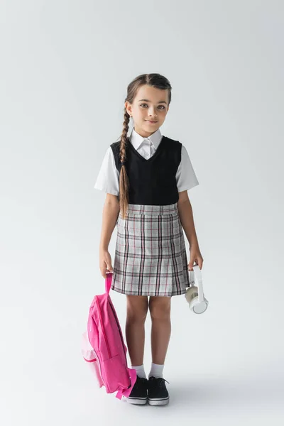 Повна довжина школярки в уніформі, стоячи з рюкзаком і тримаючи бездротові навушники на сірому — стокове фото