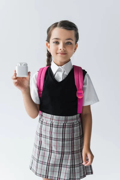 Cheerful schoolgirl in uniform standing with backpack and holding earphone case isolated on grey — Foto stock