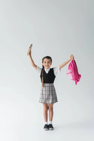Повна довжина збудженої школярки в уніформі тримає рожевий рюкзак і книгу на сірому — стокове фото