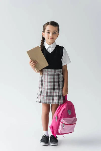 Full length of adorable schoolgirl in uniform holding pink backpack and book on grey — Stockfoto
