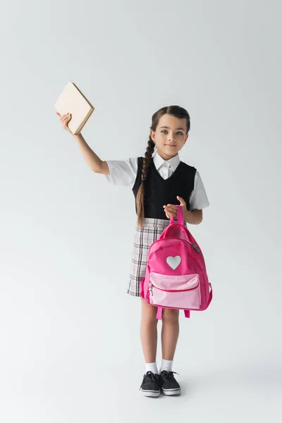 Full length of cute schoolgirl in uniform holding pink backpack and book above head on grey — Stock Photo