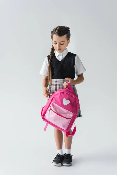 Повна довжина милої школярки в уніформі тримає рожевий рюкзак, стоячи на сірому — стокове фото