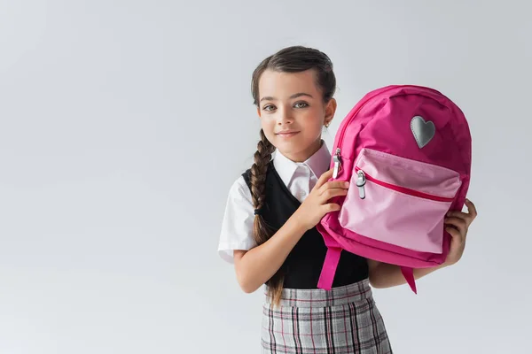 Cute schoolgirl in uniform holding pink backpack and smiling isolated on grey — стоковое фото