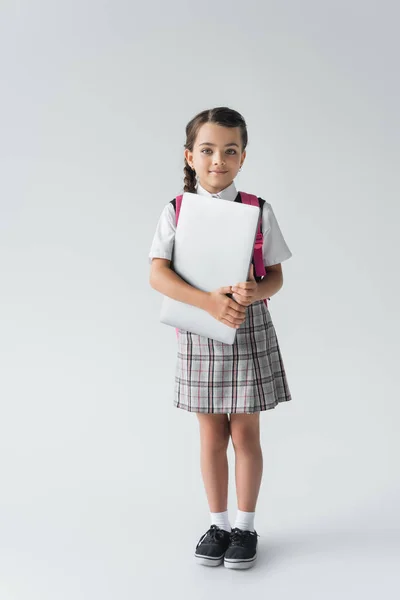 Повна довжина школярки в уніформі, що стоїть з рюкзаком і ноутбуком на сірому — стокове фото