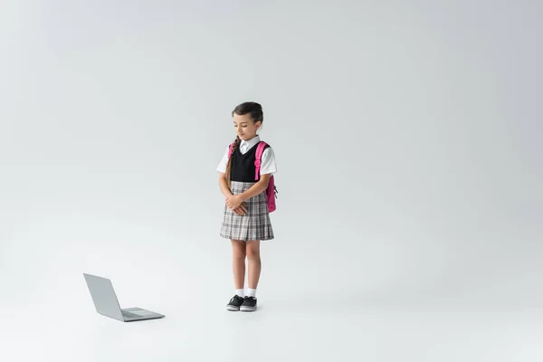 Full length of schoolgirl in uniform standing with backpack and looking at laptop on grey — Stock Photo