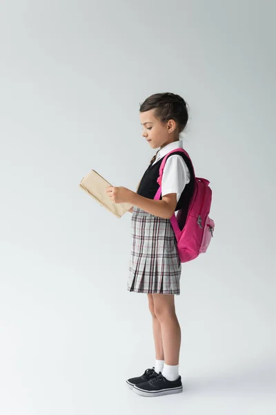 Side view of smart schoolgirl in uniform reading book and standing and reading book on grey — Foto stock