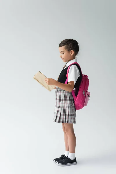 Side view of smart schoolgirl in uniform reading book and standing and reading book on grey — стоковое фото