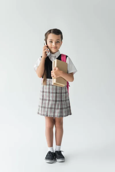 Full length of smiling schoolgirl talking on smartphone and holding books on grey — Foto stock