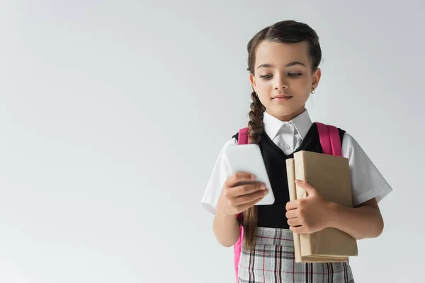 Adorable schoolgirl in uniform using smartphone and holding books isolated on grey — стоковое фото