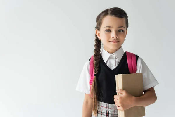Чарівна школярка в уніформі тримає книги і посміхається ізольовано на сірому — стокове фото