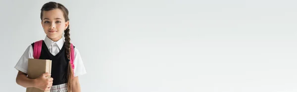 Adorable schoolgirl in uniform holding books and smiling isolated on grey, banner — Foto stock