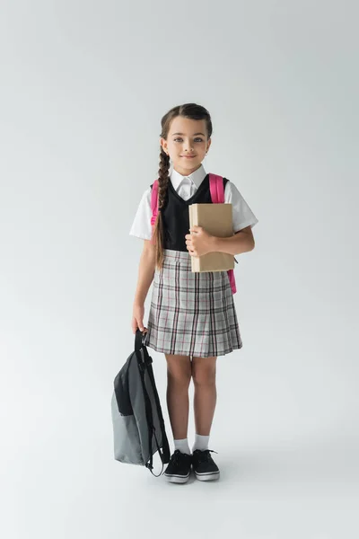 Повна довжина чарівної школярки в уніформі тримає книги і рюкзак на сірому — стокове фото