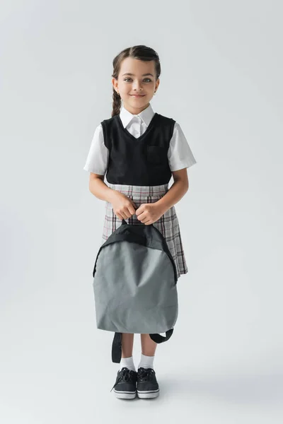 Full length of adorable and happy schoolgirl in uniform holding backpack on grey — Stockfoto