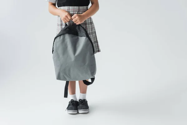 Cropped view of schoolgirl in uniform holding backpack and standing on grey — Fotografia de Stock