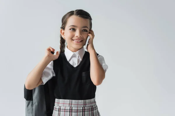 Happy schoolgirl holding backpack and talking on smartphone isolated on grey — Stock Photo