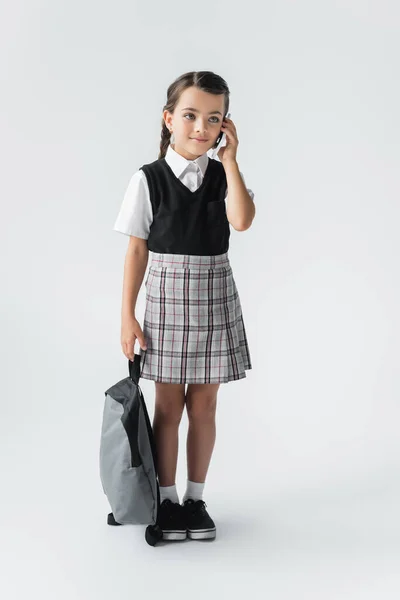 Full length of happy schoolgirl holding backpack and talking on smartphone on grey — стоковое фото