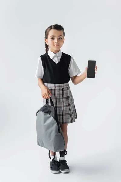 Full length of cute schoolgirl holding backpack and smartphone with blank screen on grey - foto de stock