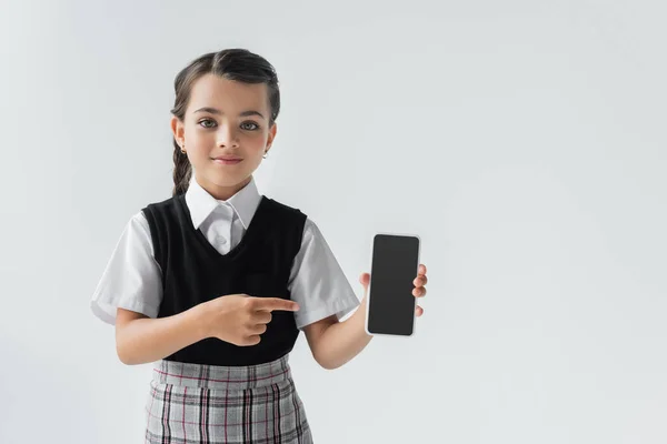 Cute schoolgirl in uniform pointing with finger at smartphone with blank screen isolated on grey — Photo de stock