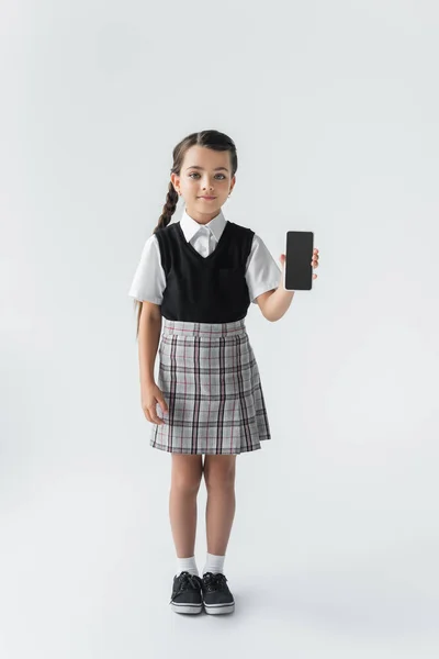 Full length of cute girl in school uniform holding smartphone with blank screen on grey — Stockfoto