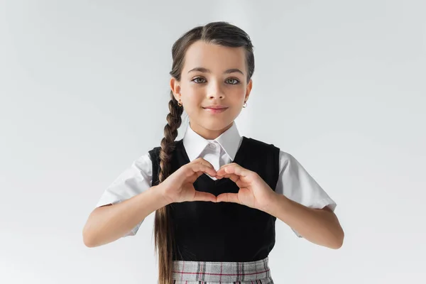 Portrait of happy schoolgirl in white shirt and vest showing heart with hands isolated on grey — Stock Photo