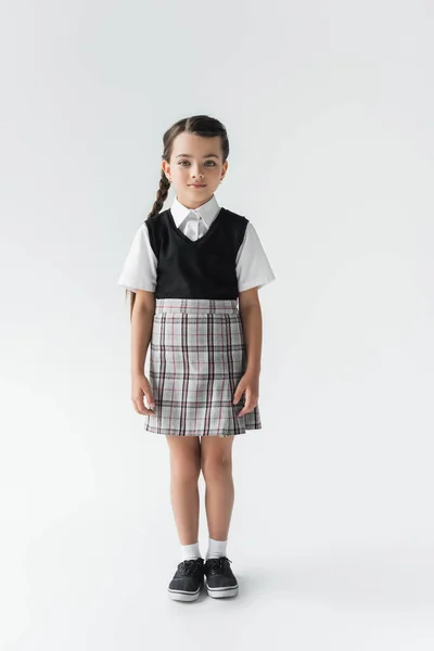 Повна довжина милої школярки в уніформі стоїть і дивиться на камеру на сірому — стокове фото