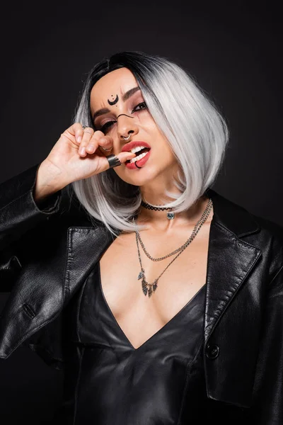 Seductora mujer con maquillaje de bruja y ceniza rubia mordiendo el pulgar aislado en negro - foto de stock