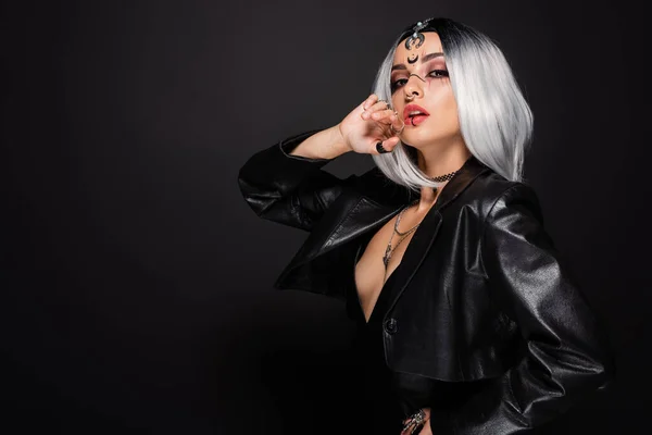 Sexy mujer en traje de bruja y maquillaje posando con la mano cerca de la cara aislado en negro - foto de stock