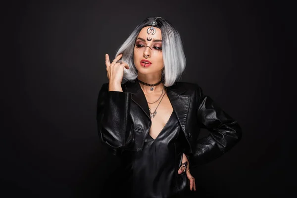 Mujer sexy en chaqueta de cuero y maquillaje estilo bruja posando con la mano en la cadera aislado en negro - foto de stock