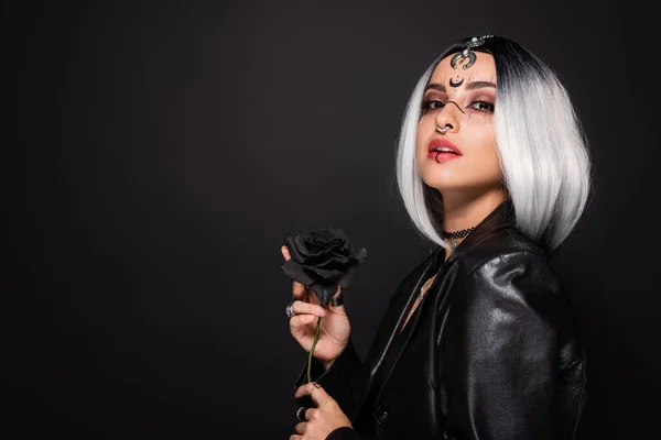 Mujer en el maquillaje de Halloween bruja y chaqueta de cuero posando con rosa oscuro aislado en negro - foto de stock