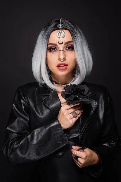 Woman in witch halloween makeup holding dark rose and looking at camera isolated on black — Stock Photo