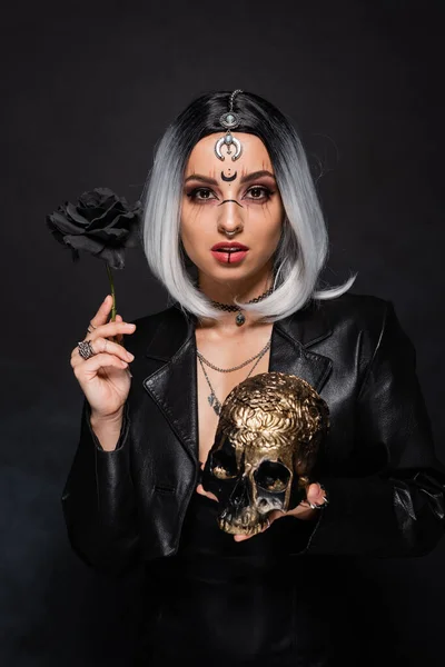 Ash blonde woman with witch makeup holding black rose and golden skull on dark background — Stock Photo