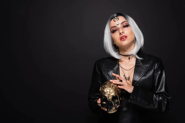 Woman in leather jacket and witch halloween makeup holding golden skull isolated on black — Stock Photo