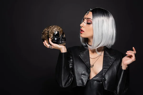 Woman in leather jacket and witch halloween makeup looking at golden skull isolated on black — Stock Photo