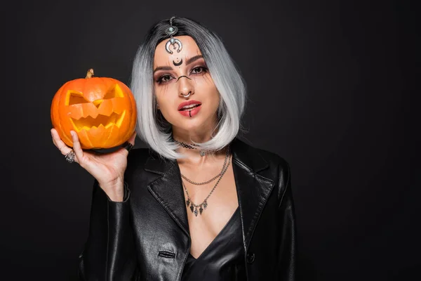 Ceniza mujer rubia en chaqueta de cuero y maquillaje de bruja celebración de calabaza tallada aislada en negro - foto de stock