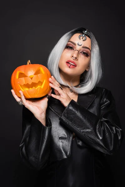 Sexy mujer de estilo bruja con calabaza tallada mirando a la cámara aislada en negro - foto de stock